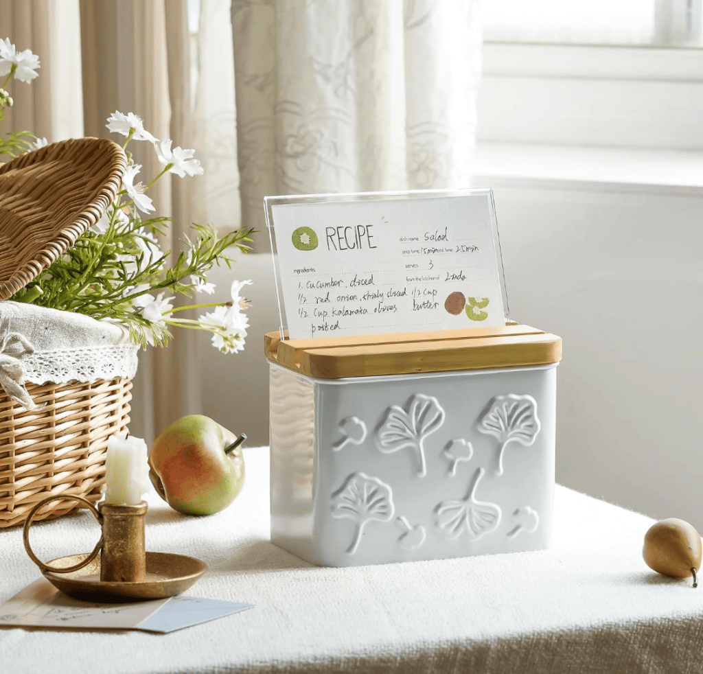 A decorative recipe box with a wooden lid, displaying a salad recipe card, positioned on a textured white tablecloth next to a candle and an apple.